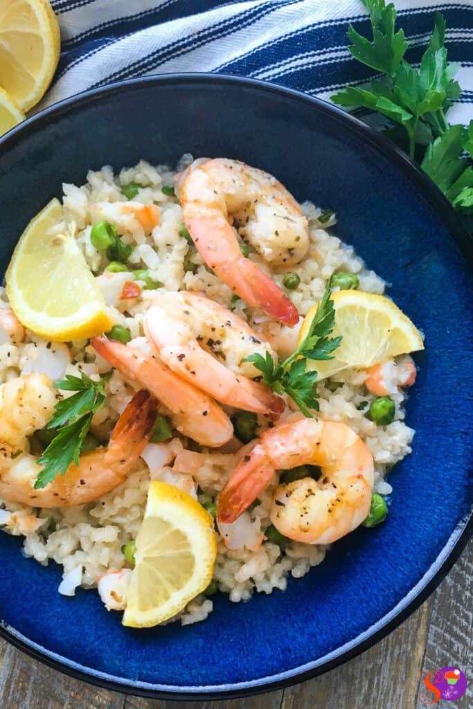 A blue bowl filled with prawn and pea risotto, garnished with lemon wedges and parsley, on a striped cloth background.