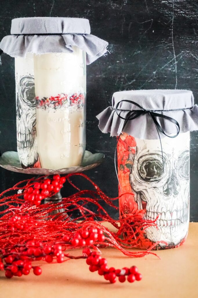 Two jars, adorned with skull illustrations, stand on a table like mysterious Skull Cookie Mix in a Jar. One jar is perched on a pedestal, surrounded by red berries and twigs, wrapped in gray fabric for an eerie yet delightful touch.