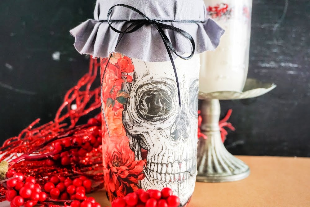 Skull Cookie Mix in a Jar 13 Decorative jar with a skull cookie mix design, gray fabric cover tied with a black ribbon. Red flowers and berries are visible around the base, adding a touch of warmth. A candle holder glows softly in the background.