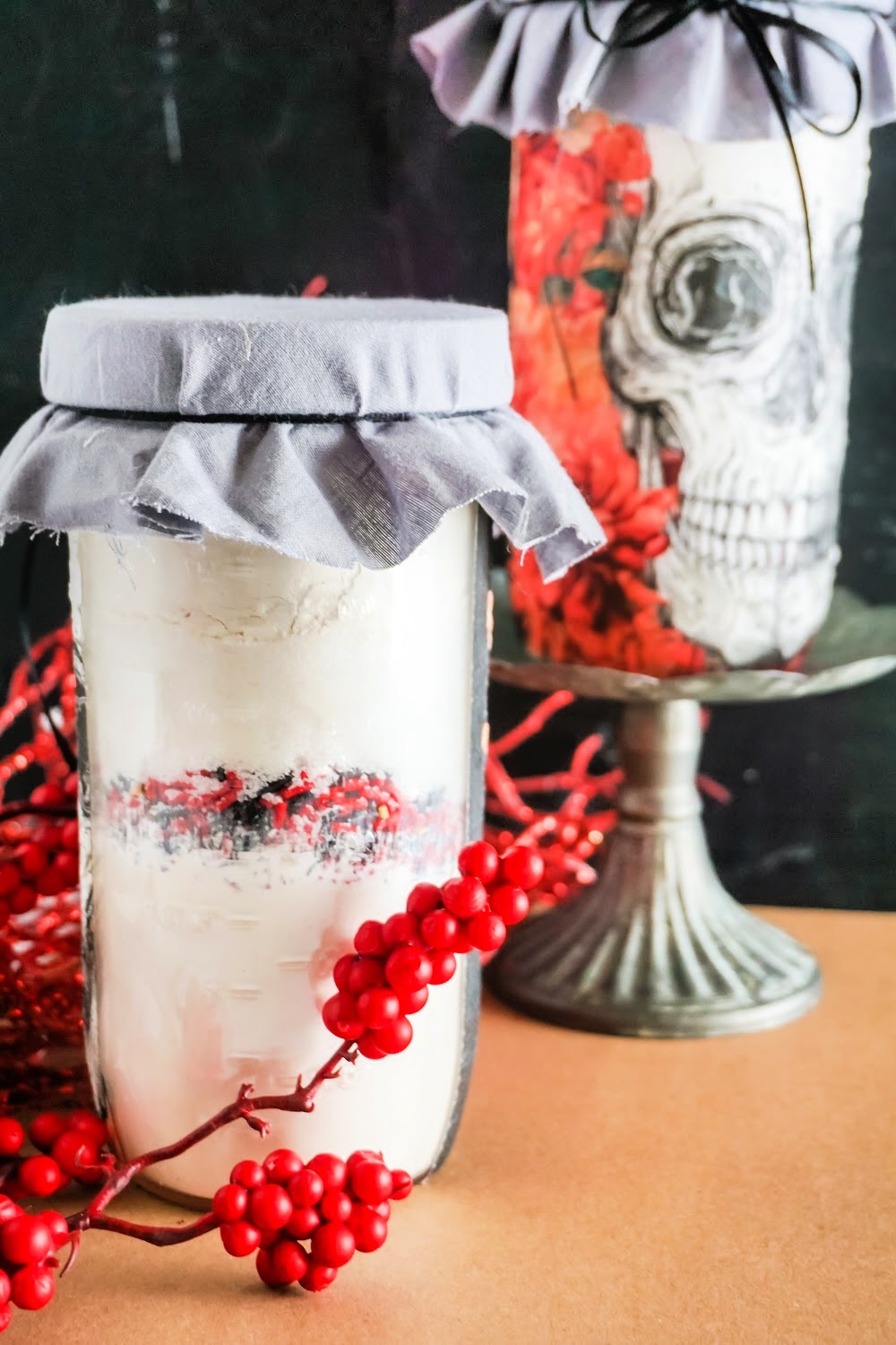 Skull Cookie Mix in a Jar 14 Two glass jars with fabric covers create a striking scene. One is filled with a white substance and red berries; the other showcases a clever skull cookie mix in a jar design. Red berries are artfully scattered, all set against a dark, moody background.