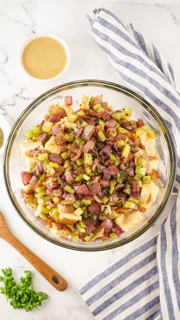 A bowl of Traeger Smoked Stuffing bread salad with red onions and herbs on a kitchen counter with a striped cloth and a wooden spoon nearby.