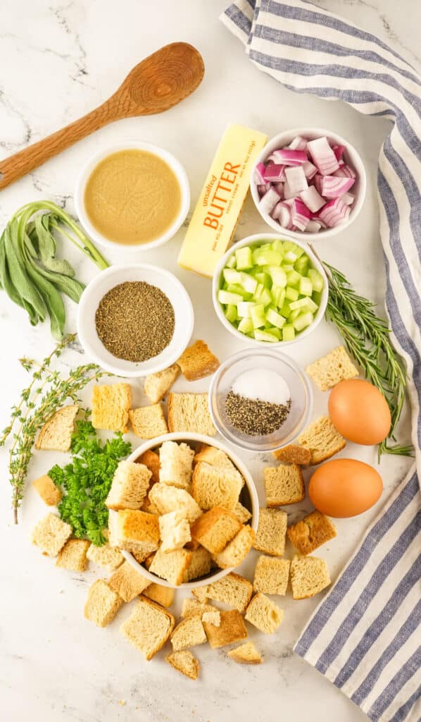 Ingredients for cooking arrayed on a kitchen counter, including bread cubes, herbs, eggs, diced vegetables, and seasonings for Traeger Smoked Stuffing.