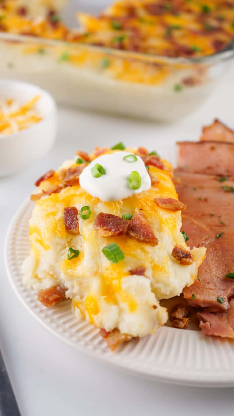 A plate showcasing sliced ham alongside loaded mashed potatoes, topped with cheese, bacon bits, sour cream, and chopped green onions. A dish of shredded cheese is in the background. This hearty meal highlights the irresistible charm of Loaded Baked Mashed Potato delight.