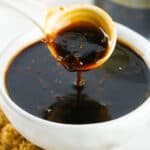 Spoon pouring thick teriyaki sauce into a white bowl, with granulated sugar and ginger root visible in the background.