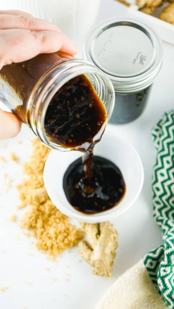 A person pours dark homemade teriyaki sauce from a glass jar into a small white bowl, with brown sugar and a cloth nearby on a white surface.