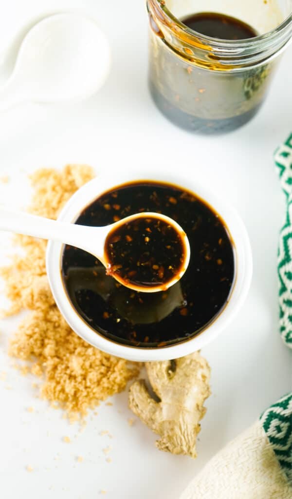 A white spoon lifting homemade Teriyaki sauce from a small bowl surrounded by ginger, a jar, and a napkin on a white surface.