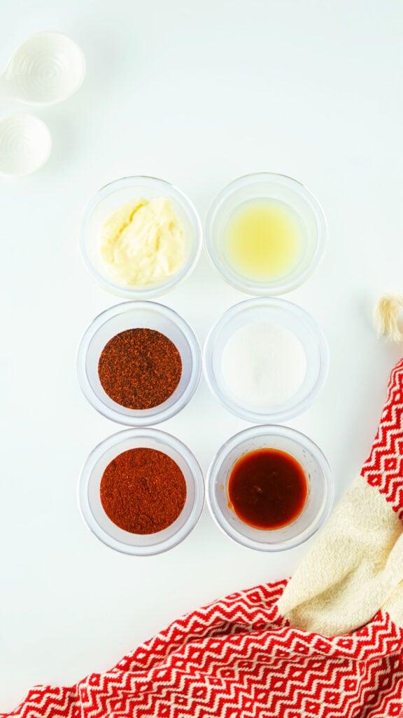 Overhead view of various ingredients in small bowls, including spices and homemade Chipotle Mayo, on a white surface with a red and white towel on the side.