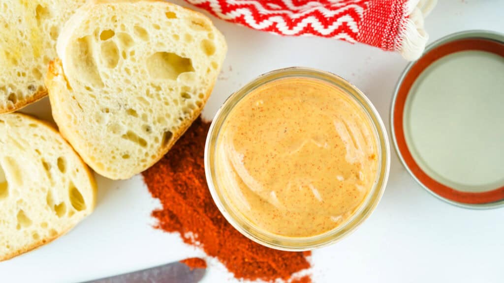 Overhead view of a glass bowl of homemade chipotle mayo, sliced baguette, a dish of cream, paprika, and a red and white napkin on a white surface.