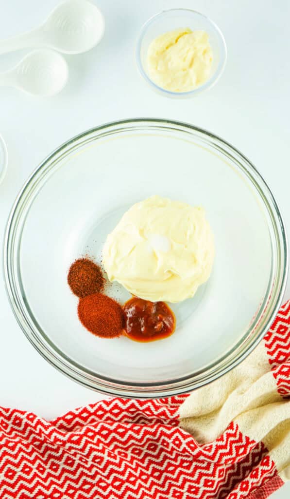 A bowl containing homemade chipotle mayo, set on a white surface with measuring spoons and a red patterned cloth nearby.