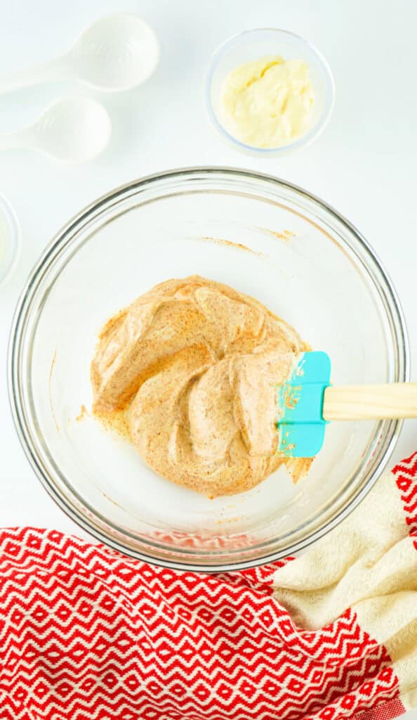 A bowl of mixed batter with a blue spatula on a red and white cloth, surrounded by measuring spoons and a small bowl of chipotle mayo.