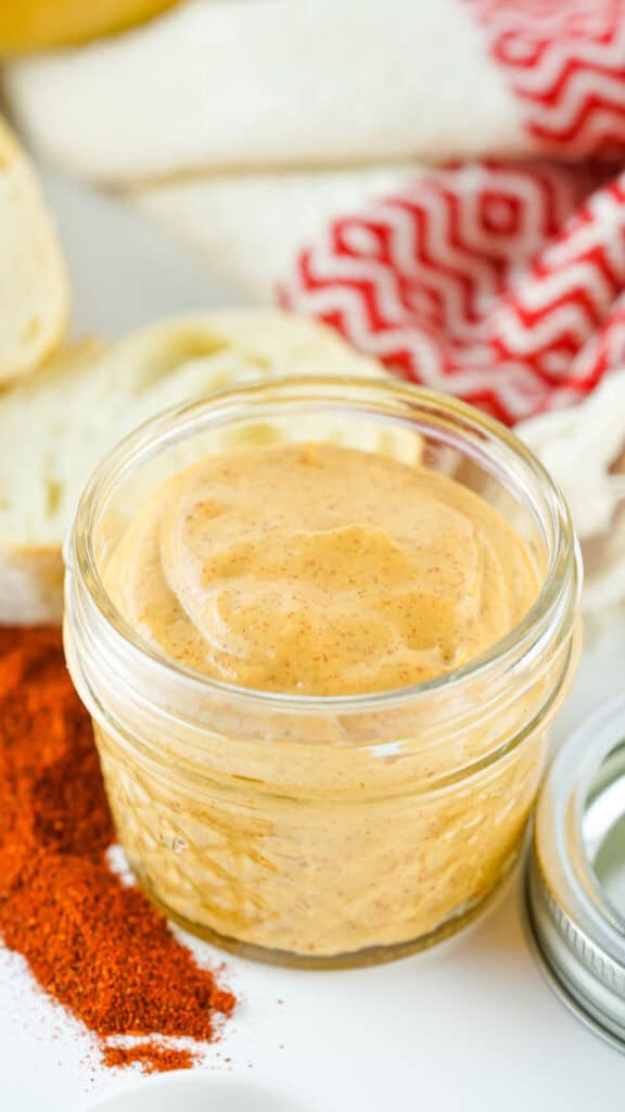 A close-up of a jar of homemade chipotle mayo with spices visible, accompanied by a checkered red and white cloth and fresh ingredients in the background.