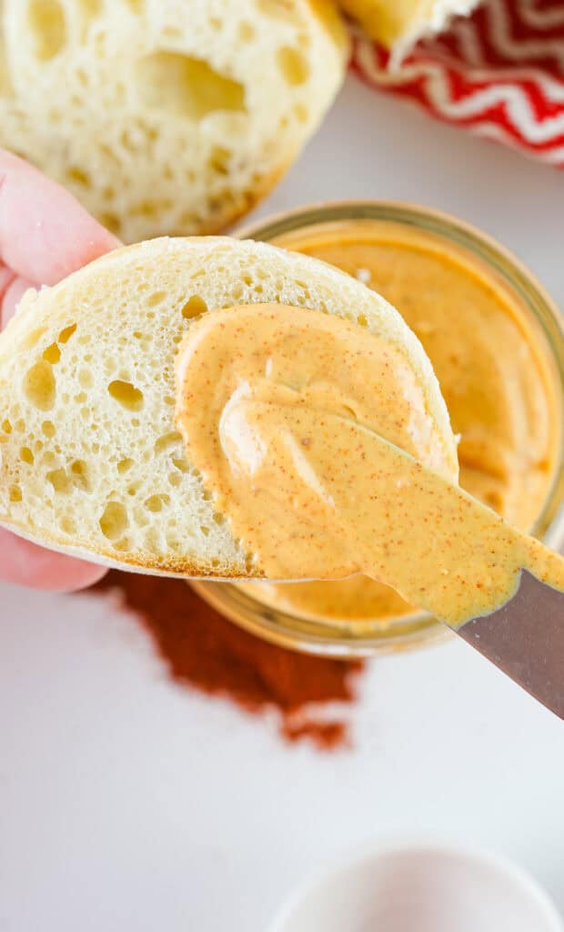 A person spreading homemade chipotle mayo on a slice of bread, with more sauce in a jar and spices scattered in the background.