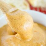 Close-up of a wooden spoon dripping with creamy, spiced Chipotle Mayo, served in a white bowl with grated cheese visible in the background.