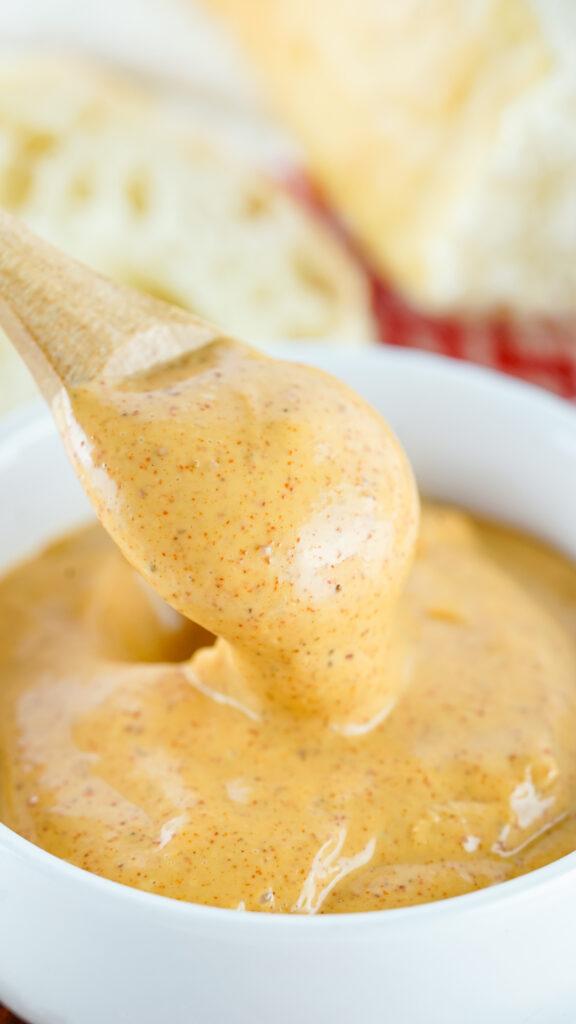 Close-up of a wooden spoon dripping with creamy, spiced Chipotle Mayo, served in a white bowl with grated cheese visible in the background.