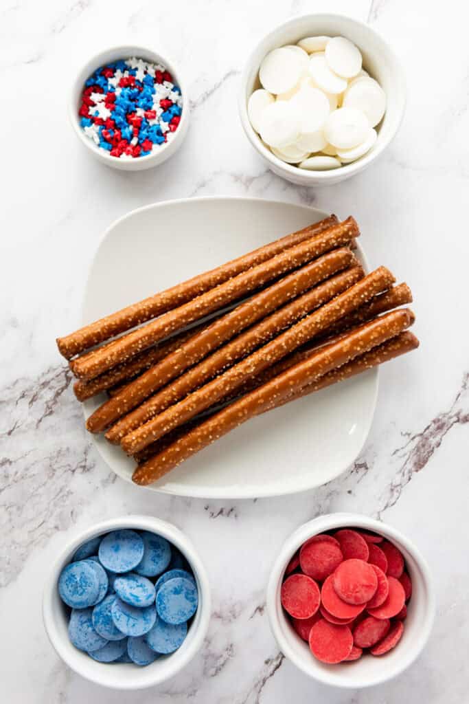 A white plate with pretzel rods surrounded by small bowls containing red, blue, and white candy melts and red, white, and blue star-shaped sprinkles on a marble surface create festive treats perfect for easy party snacks.