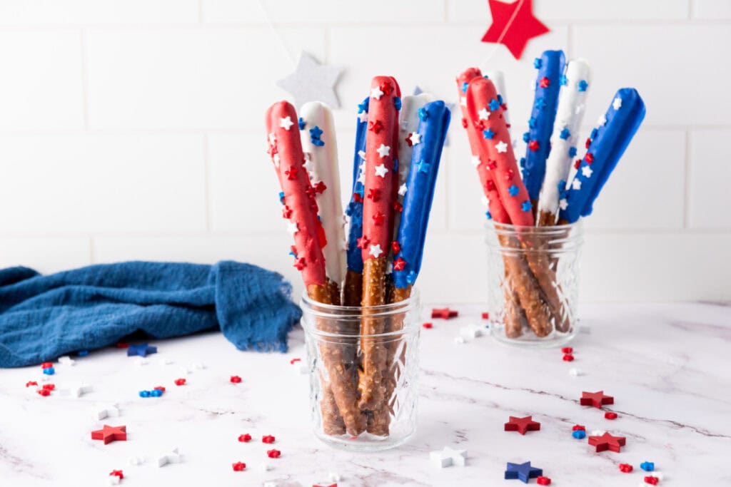 Glass jars filled with candy-coated pretzel rods in blue, red, and white chocolate, decorated with star-shaped sprinkles. Scattered sprinkles adorn the white surface, with a blue cloth in the background. Perfect Fourth of July snacks that add a festive touch to your patriotic celebrations.