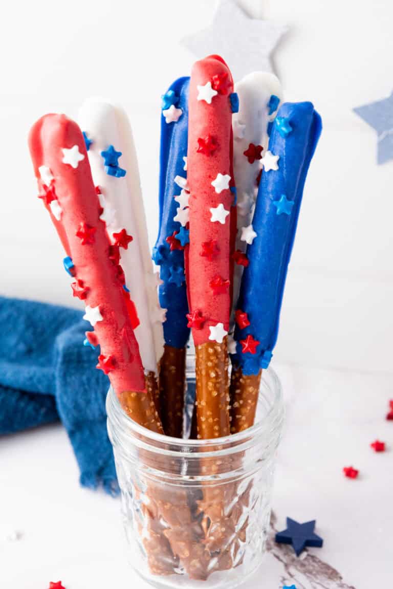 A glass jar holds Firecracker Pretzel Rods dipped in red, white, and blue chocolate, decorated with star-shaped sprinkles—perfect for easy party snacks.