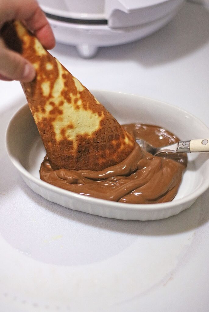 A hand dips a folded crepe into a dish of thick chocolate sauce with a spoon on the side, reminiscent of indulgent flavors found in Ben & Jerry’s Copycat No Churn Homemade Americone Dream Ice Cream.