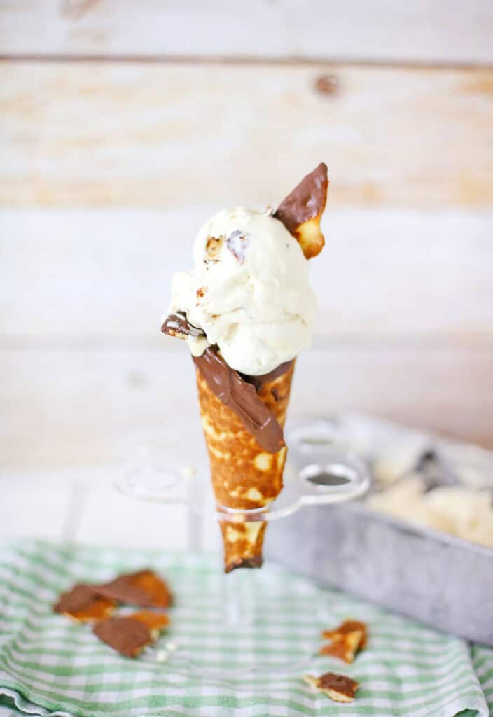 A waffle cone filled with two scoops of Ben & Jerry’s Americone Dream ice cream, topped with chocolate pieces, stands upright on a metal holder. Crumbled cone pieces and a green checkered cloth are in the foreground.