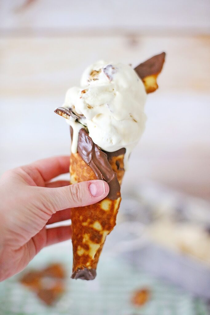 A hand holds a chocolate-dipped waffle cone filled with creamy vanilla ice cream reminiscent of Ben & Jerry’s Copycat No Churn Homemade Americone Dream Ice Cream, elegantly topped with a chocolate piece.