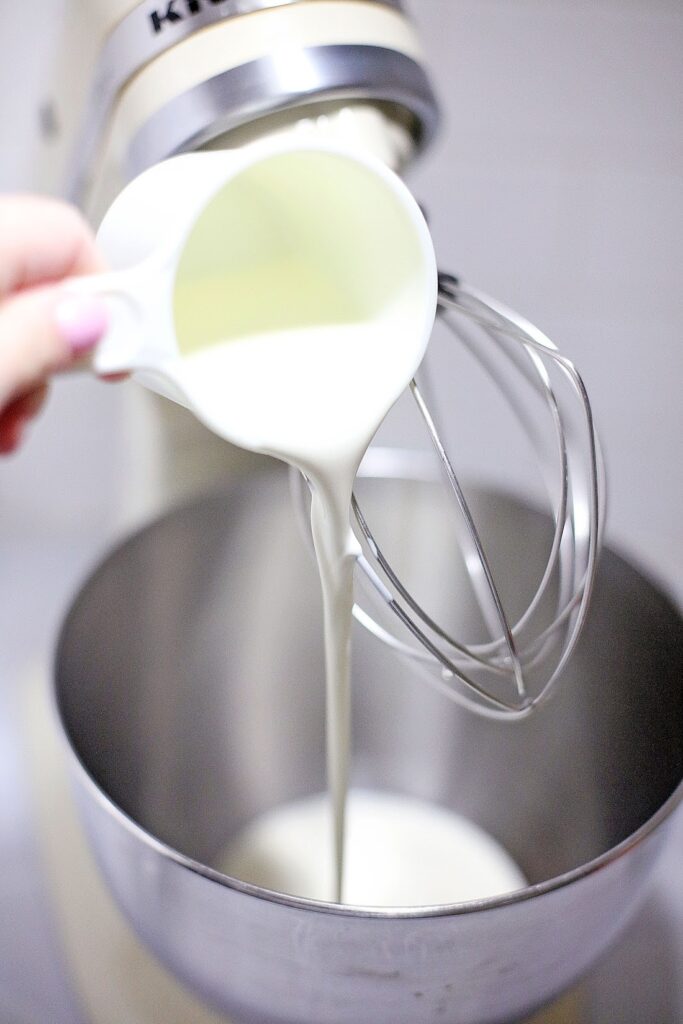 A person pouring cream into a stand mixer bowl with a whisk attachment, preparing for a batch of Ben & Jerry’s Copycat No Churn Homemade Americone Dream Ice Cream.