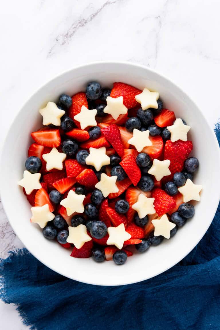 A bowl filled with cut strawberries, blueberries, and star-shaped pieces of white fruit rests on a dark blue cloth, creating a Star Spangled Fruit Salad.