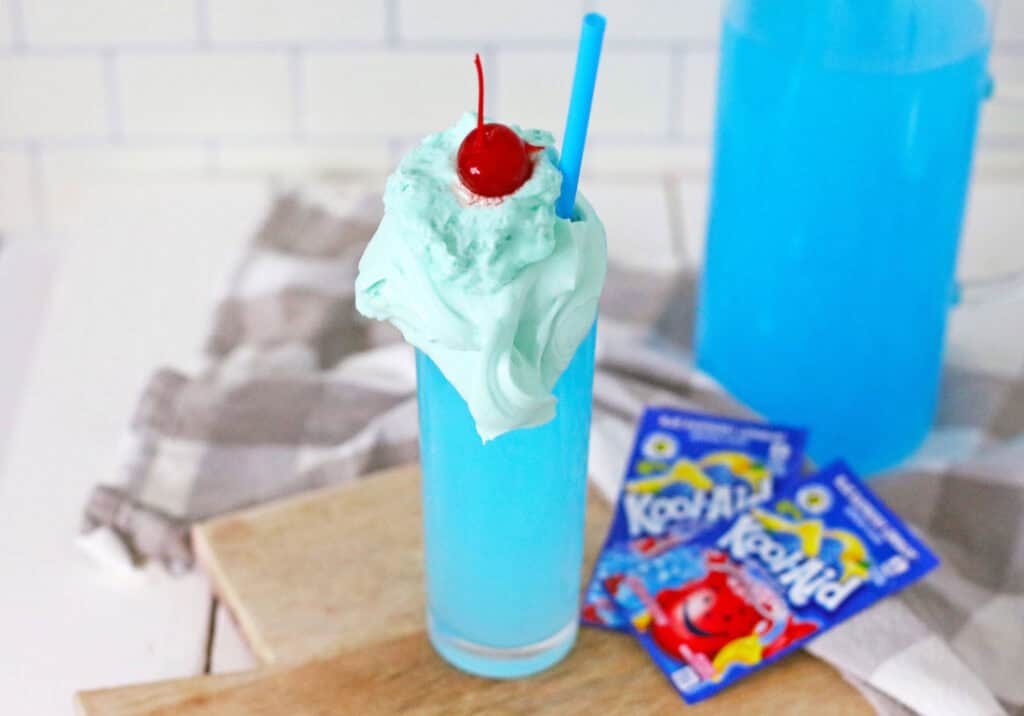 A tall glass of blue Whipped Kool-Aid topped with whipped cream, a cherry, and a blue straw, placed on a wooden board with Kool-Aid packets in the background.