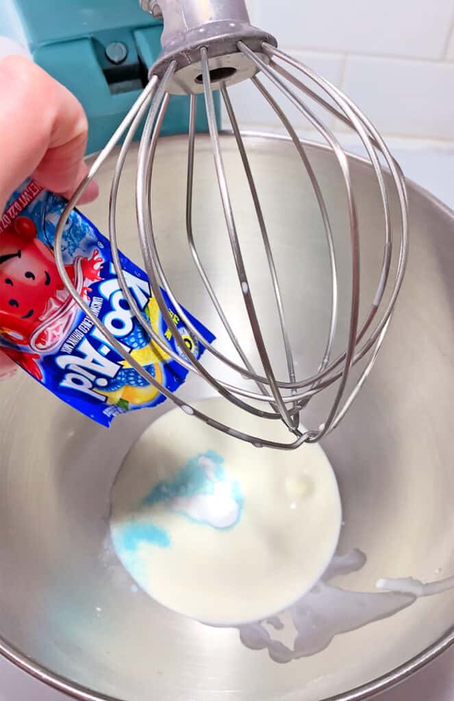 Pouring packets of blue Kool-Aid powder into a mixing bowl containing a white liquid, with a whisk attachment in view, creates the perfect base for a delightful Whipped Kool-Aid drink.