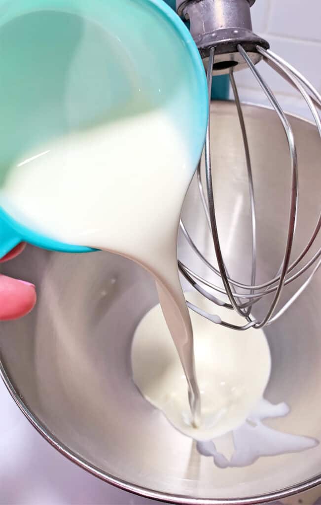 Milk being poured from a turquoise bowl into a metal mixing bowl with a whisk attachment, ready to craft the perfect whipped drink.