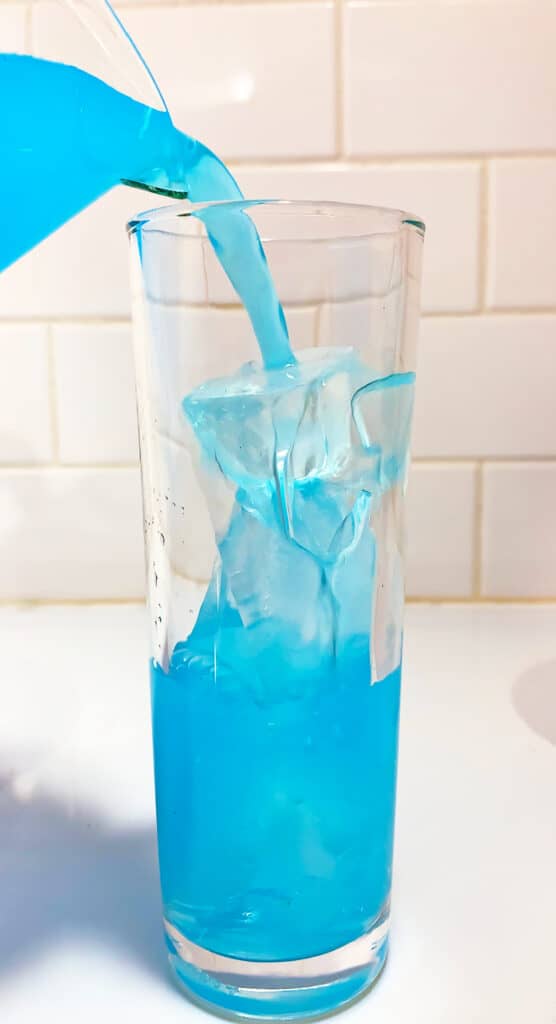 A whipped Kool-Aid beverage is being poured into a tall glass filled with ice cubes against a white tiled background.