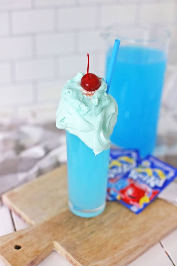 A tall glass filled with a whipped Kool-Aid blue drink topped with whipped cream and a cherry, along with a blue straw, on a wooden board. A pitcher of the same whipped drink and pouches of Kool-Aid are in the background.