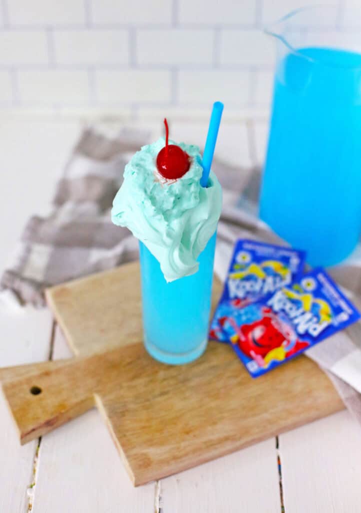 A tall glass with blue whipped Kool-Aid, topped with blue whipped cream and a red cherry, positioned on a wooden cutting board. Blue Kool-Aid packets and a pitcher of the vibrant whipped drink are in the background.