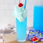 A tall glass filled with Whipped Kool-Aid topped with whipped cream and a cherry, served with a blue straw. Two packets of Kool-Aid are placed on a wooden board next to the glass.