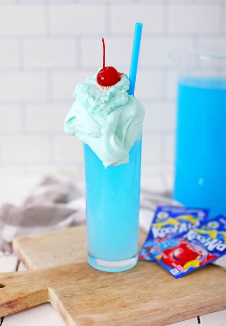 A tall glass filled with Whipped Kool-Aid topped with whipped cream and a cherry, served with a blue straw. Two packets of Kool-Aid are placed on a wooden board next to the glass.