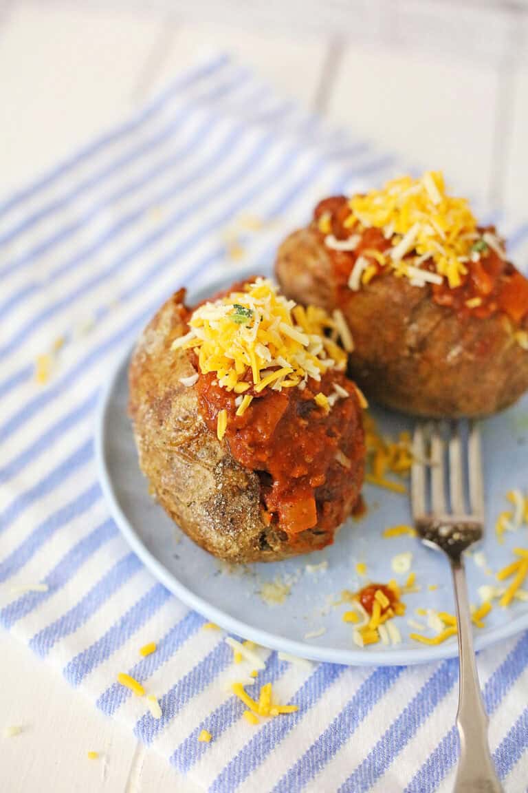 Two chili cheese baked potatoes topped with shredded cheese are served on a blue plate with a fork, placed on a blue and white striped cloth.