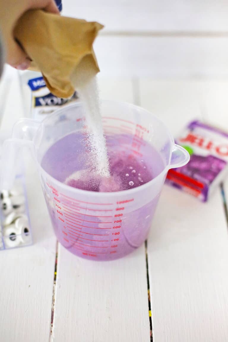 Six purple gelatin cups, dubbed Creepy Monster Eye Jello Shots, with candy eyes on top, are artfully arranged on a fringed orange mat resting on a white surface.