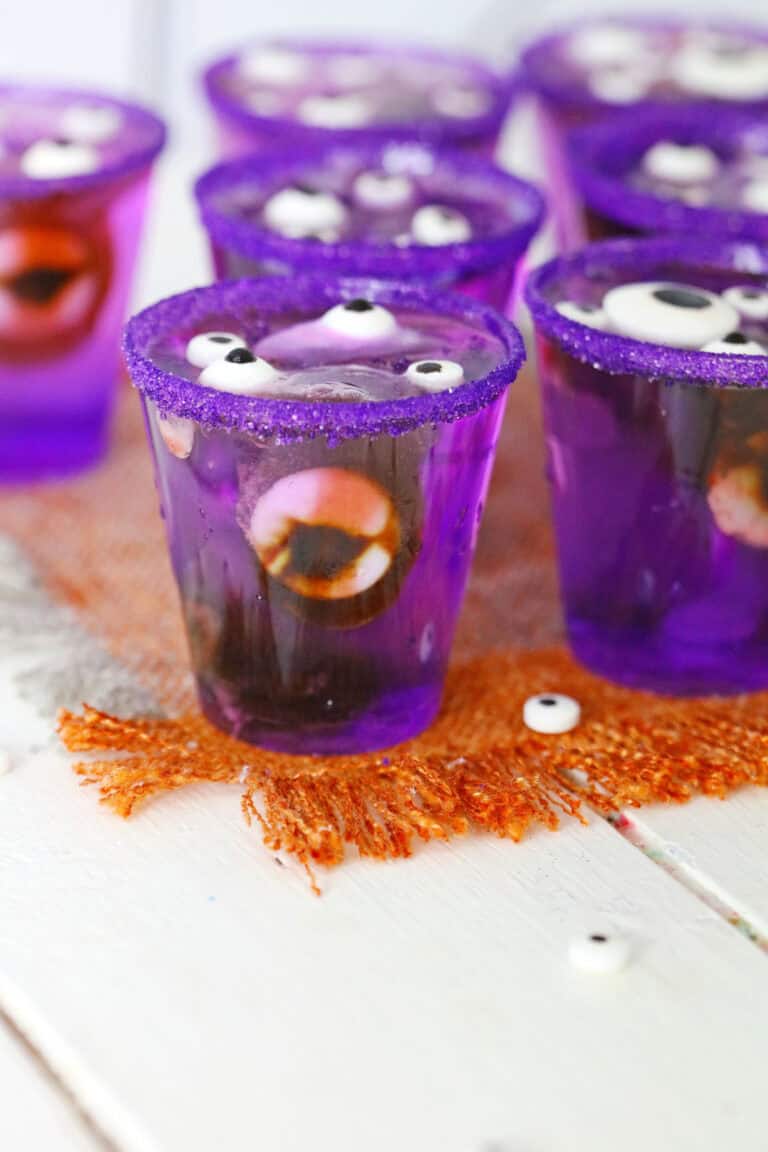 Purple gelatin cups, dubbed Monster Eye Jello Shots, are decorated with candy eyes and purple sugar rims. They sit on an orange shredded decoration on a white wooden surface.