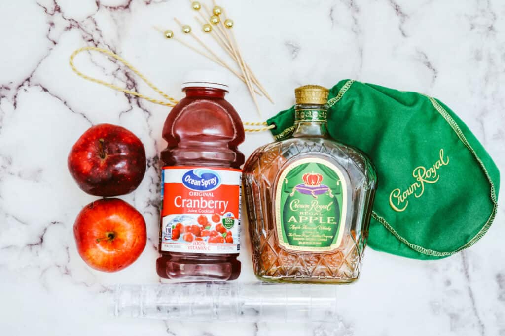 Image of ingredients for a Crown Cran Apple Shooter, featuring a bottle of Crown Royal Apple whisky, a bottle of Ocean Spray cranberry juice, two apples, a green pouch, and some garnish sticks on a marble surface.