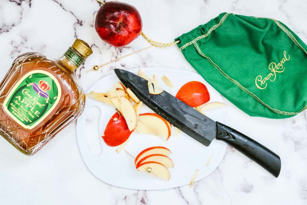 Bottle of Crown Royal Apple, a red apple, a green drawstring bag, and a knife on a cutting board with apple slices on a marble surface, perfect for preparing your Crown Cran Apple Shooter.