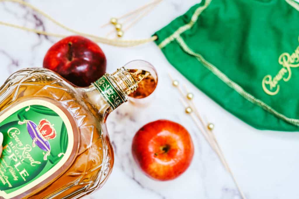 A bottle of Crown Royal Regal Apple whiskey next to apples and a green Crown Royal bag on a marble surface, perfect for crafting the ultimate Crown Cran Apple Shooter.