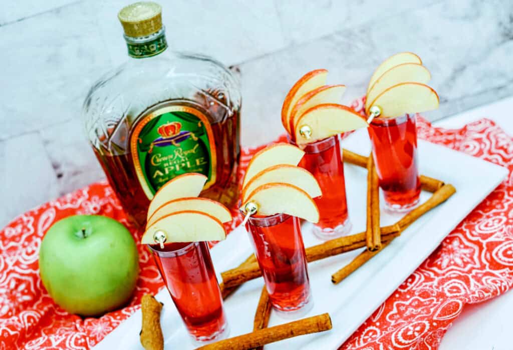 A bottle of Crown Royal Apple whiskey beside four apple-garnished red shot glasses, a green apple, and cinnamon sticks on a white tray with a red patterned cloth underneath create the perfect setup for a delightful Crown Cran Apple Shooter experience.