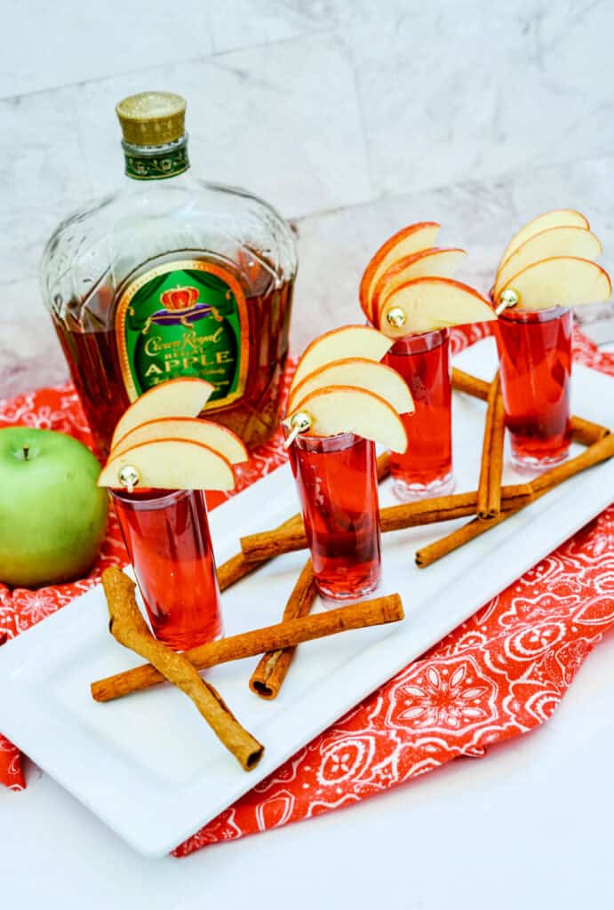 A bottle of Crown Royal Apple whiskey, an apple, and four Crown Cran Apple Shooter cocktails in shot glasses garnished with apple slices and cinnamon sticks are placed on a white tray and red patterned cloth.