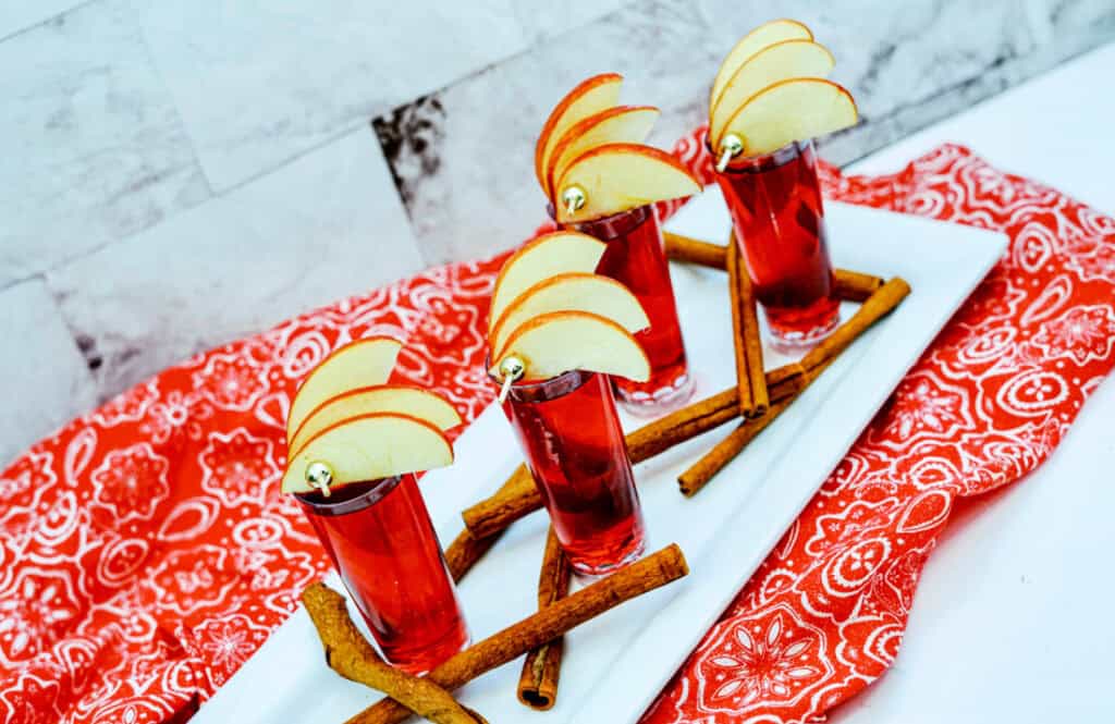 Four red Crown Cran Apple Shooter cocktails garnished with apple slices and cinnamon sticks are served on a white tray placed on a red patterned cloth.