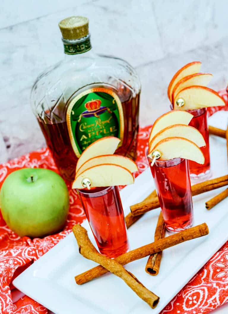 Three red Crown Cran Apple Shooters with apple slices and a bottle of Crown Royal Apple beside a green apple and cinnamon sticks on a white plate.