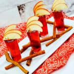 Four Crown Cran Apple Shooters in shot glasses garnished with apple slices, set on a white rectangular tray with cinnamon sticks and a red patterned cloth underneath.