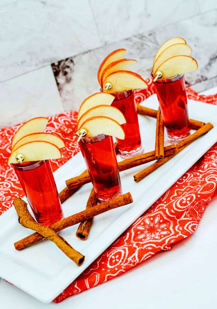 Four Crown Cran Apple Shooters in shot glasses garnished with apple slices, set on a white rectangular tray with cinnamon sticks and a red patterned cloth underneath.