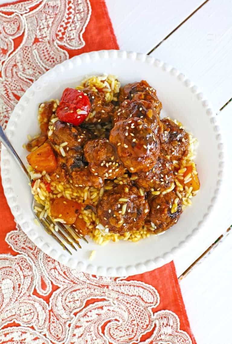 A white plate containing sweet and sour meatballs covered in sauce, mixed with vegetables, and served over rice. A fork rests on the plate, and the setting includes a red and white decorative placemat.