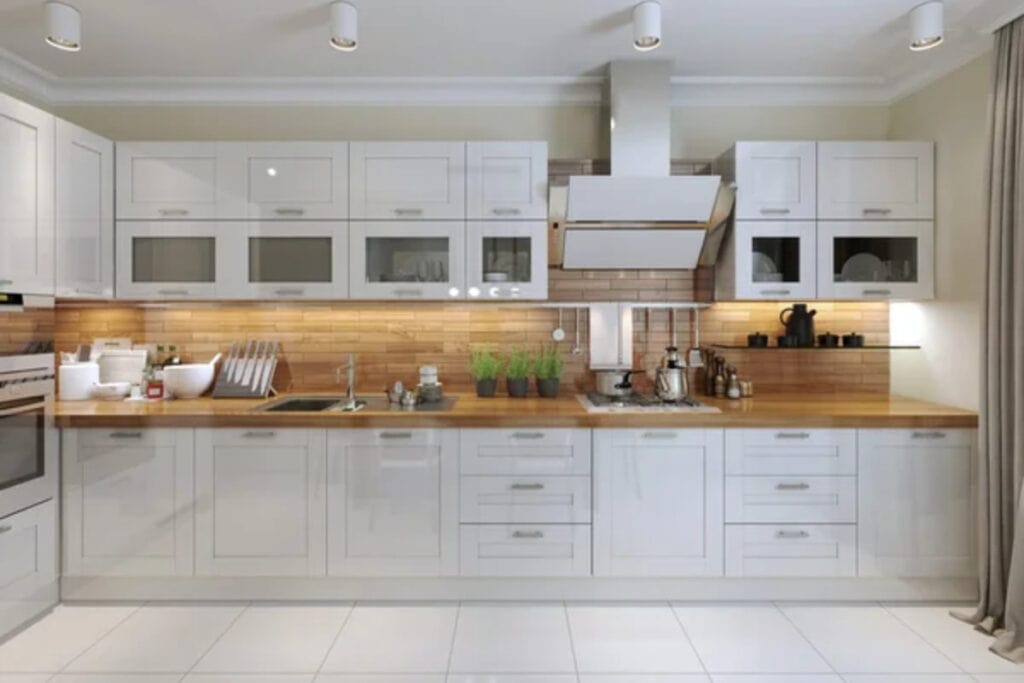 A modern kitchen with white cabinets, a wooden countertop, and stainless steel appliances, including a stove and range hood. The backsplash is tiled in a beige color, and various kitchen items are on the counter.