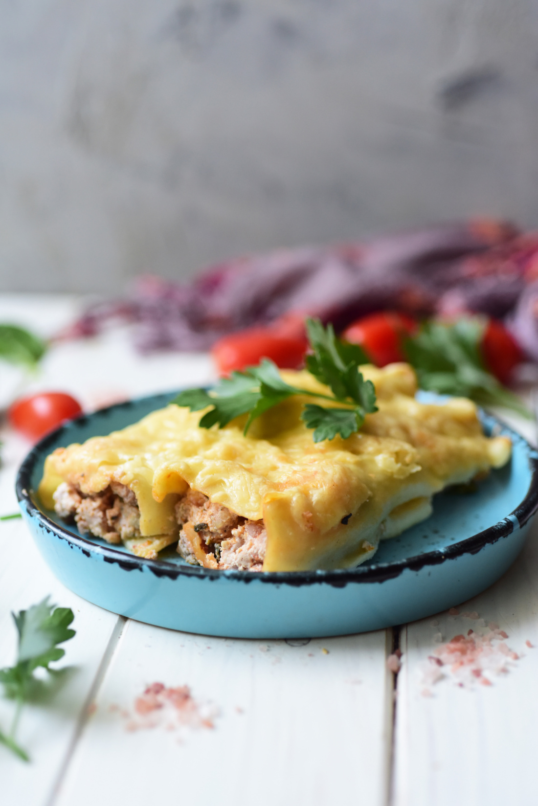 A plate of cheesy Italian Sausage and Ricotta Cannelloni topped with parsley on a blue dish. Tomatoes and herbs are in the background, adding a touch of rustic charm.