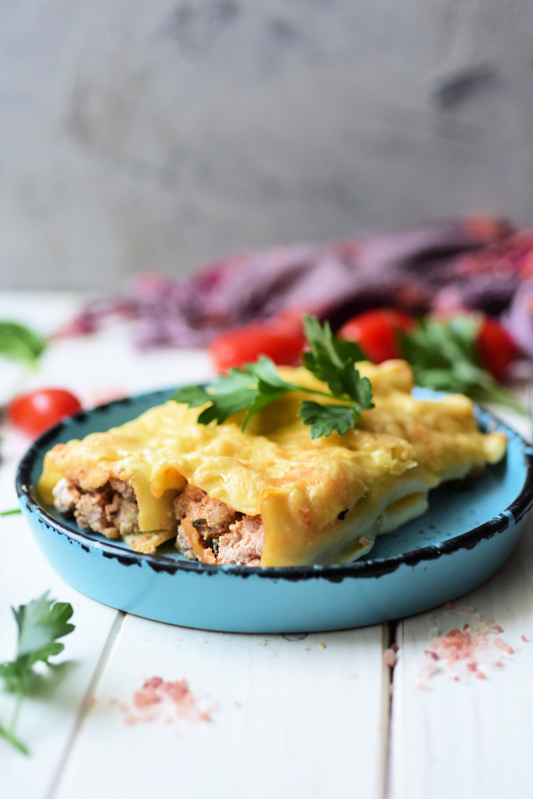 A plate with two Italian Sausage and Ricotta Cannelloni pieces topped with melted cheese and garnished with fresh parsley, placed on a white wooden table with scattered greens and cherry tomatoes in the background.